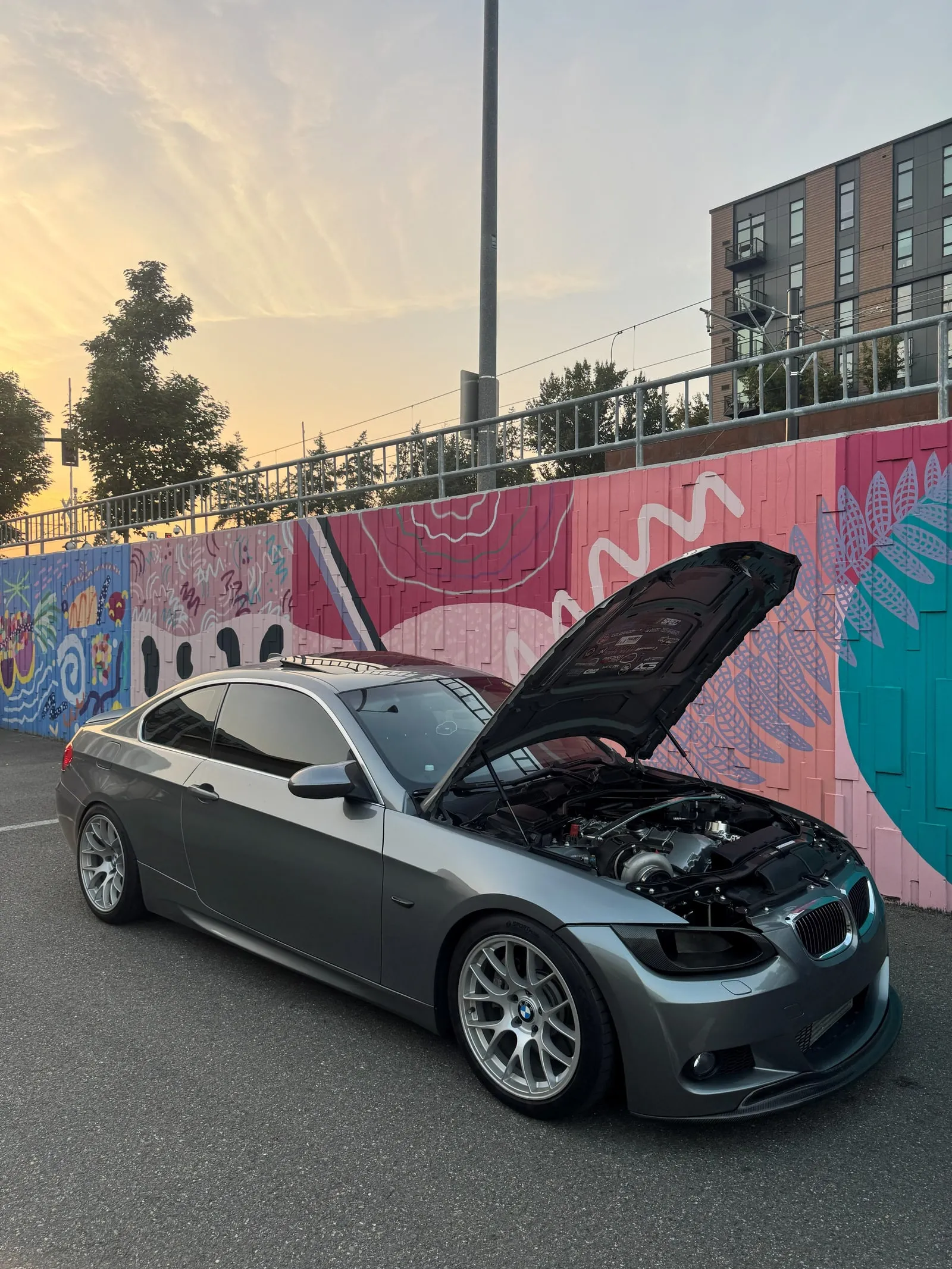 Silver E92 335i single-turbo build with the hood up, in front of a colorful mural at sunset