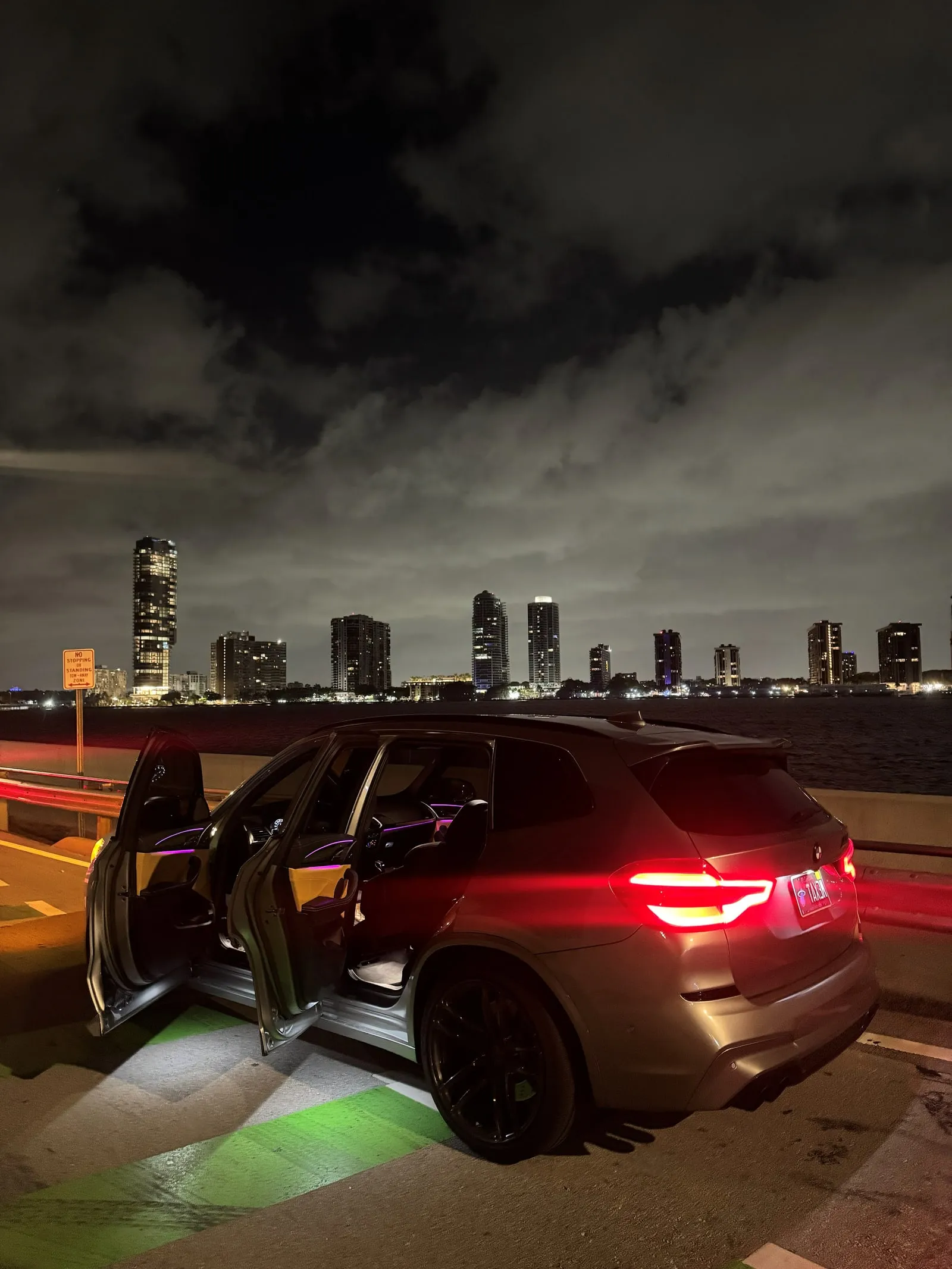 BMW X3M Competition at night with doors open and ambient lighting, city skyline behind